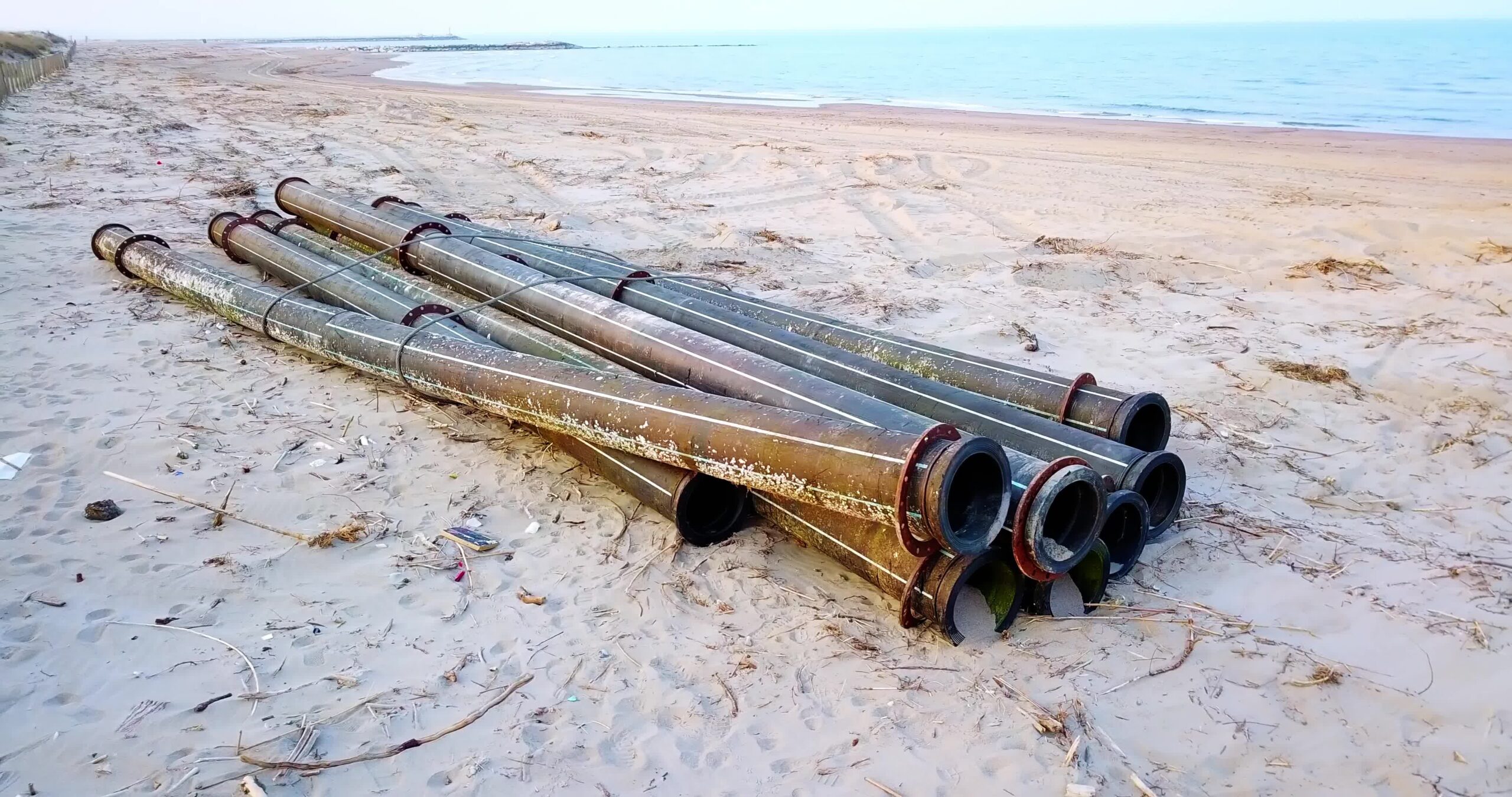 Black metal pipes lie against fence on beach with rubbish