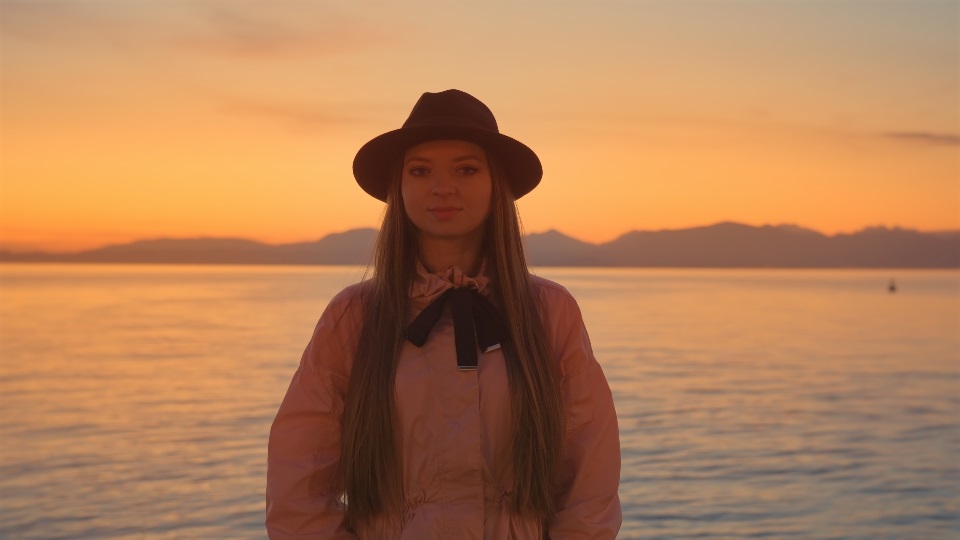 Young woman in black hat stands against Lake Garda at sunset