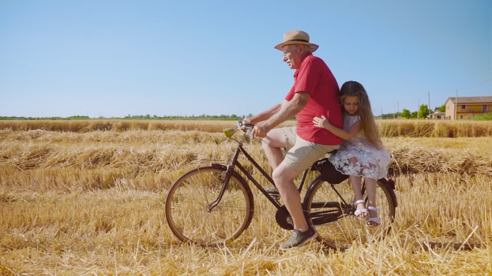 Grandfather carries granddaughter on bicycle past field