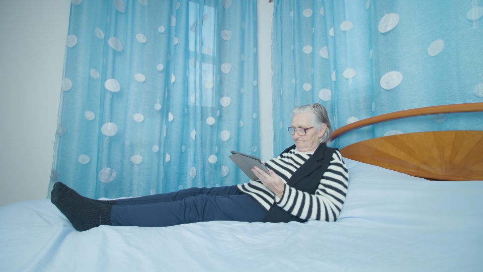 Old grey-haired woman reads news via tablet lying on bed