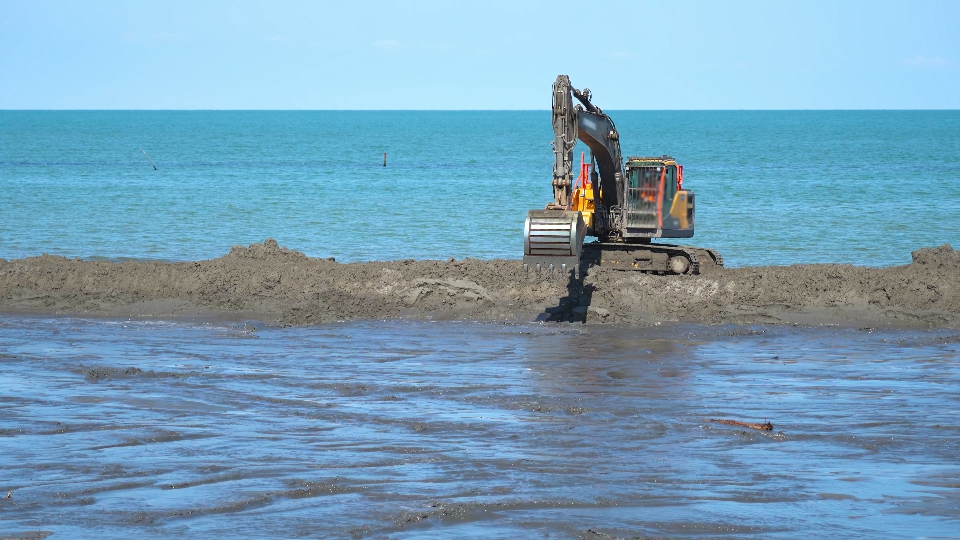 An excavator moves on the sand dune in front of the blue sea