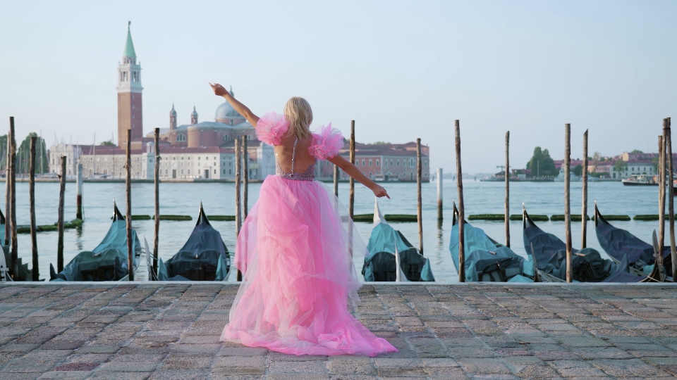 Woman with heels and pink dress walks past moored gondolas in Venice